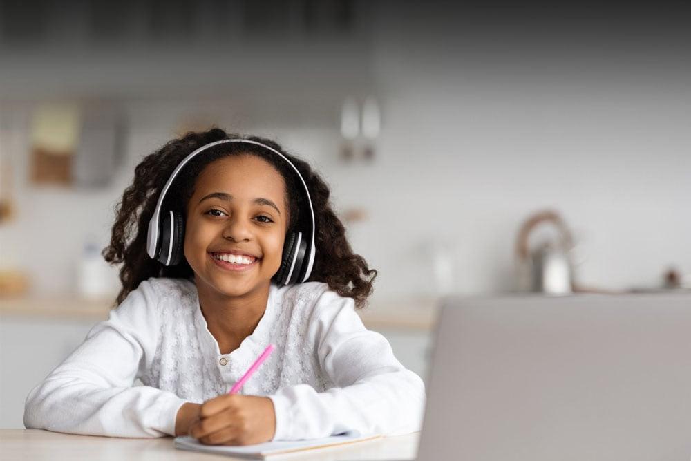 Girl in a white shirt looking at the camera smiling while writing in her school journal taking an online class at Inspire Connections Academy. 